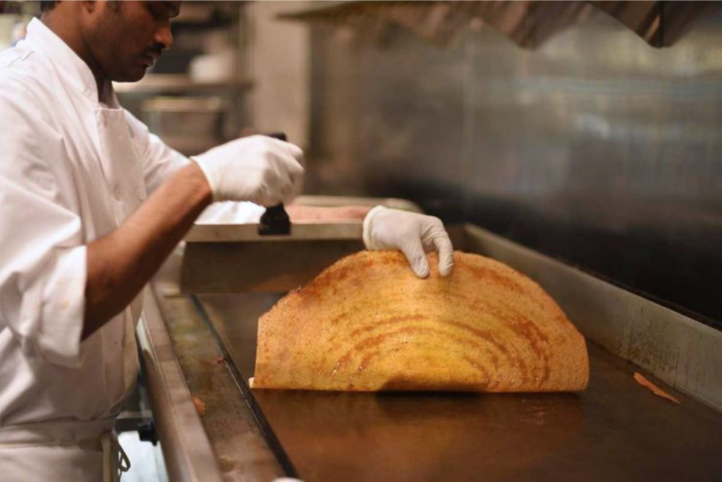 A cook spreading dosa batter across a hot griddle.